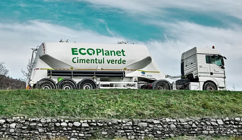 Bílá nákladní s přívězem pro transport cementu na trávnaté svahu.